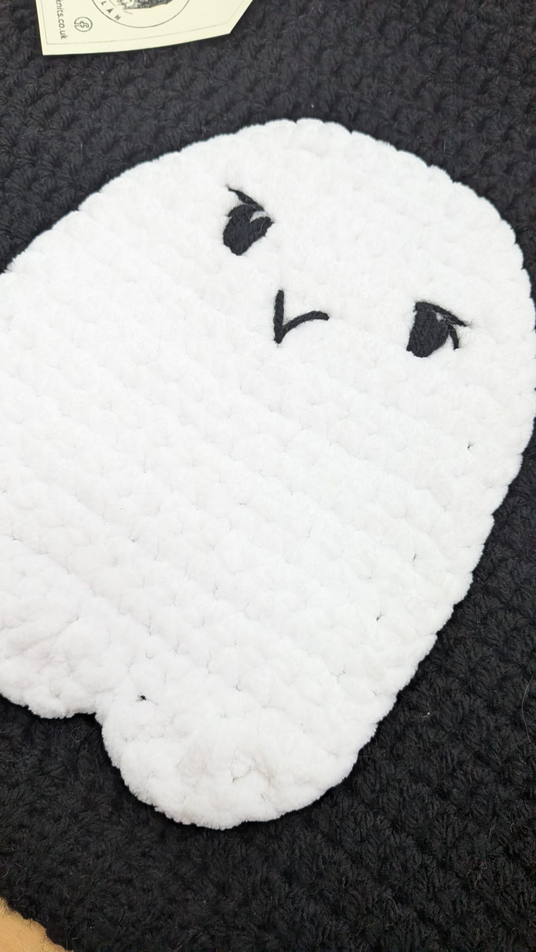 A white crocheted ghost-shaped cushion with embroidered black eyes and mouth, resting on a dark charcoal knit blanket.
