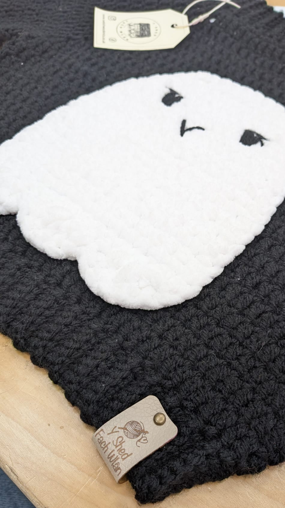 A close-up of a black knit garment, likely a sweater or hat, featuring a large white ghost applique with embroidered eyes and mouth. A small fabric tag is visible near the bottom edge, and the item is laid on a light wood surface.