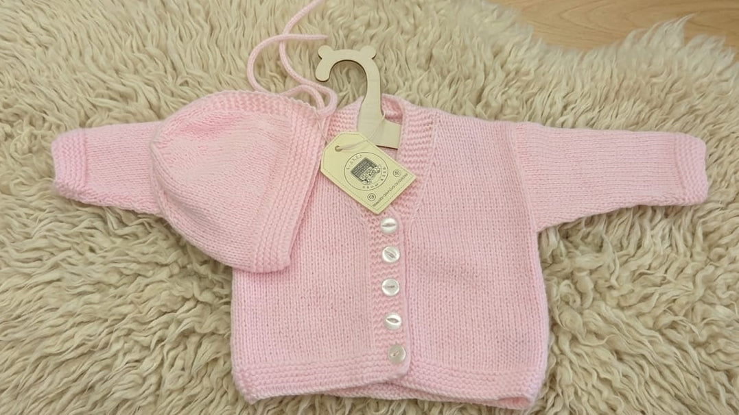 A light pink hand-knit baby cardigan and matching bonnet set with white buttons, displayed on a cream-colored shag rug.
