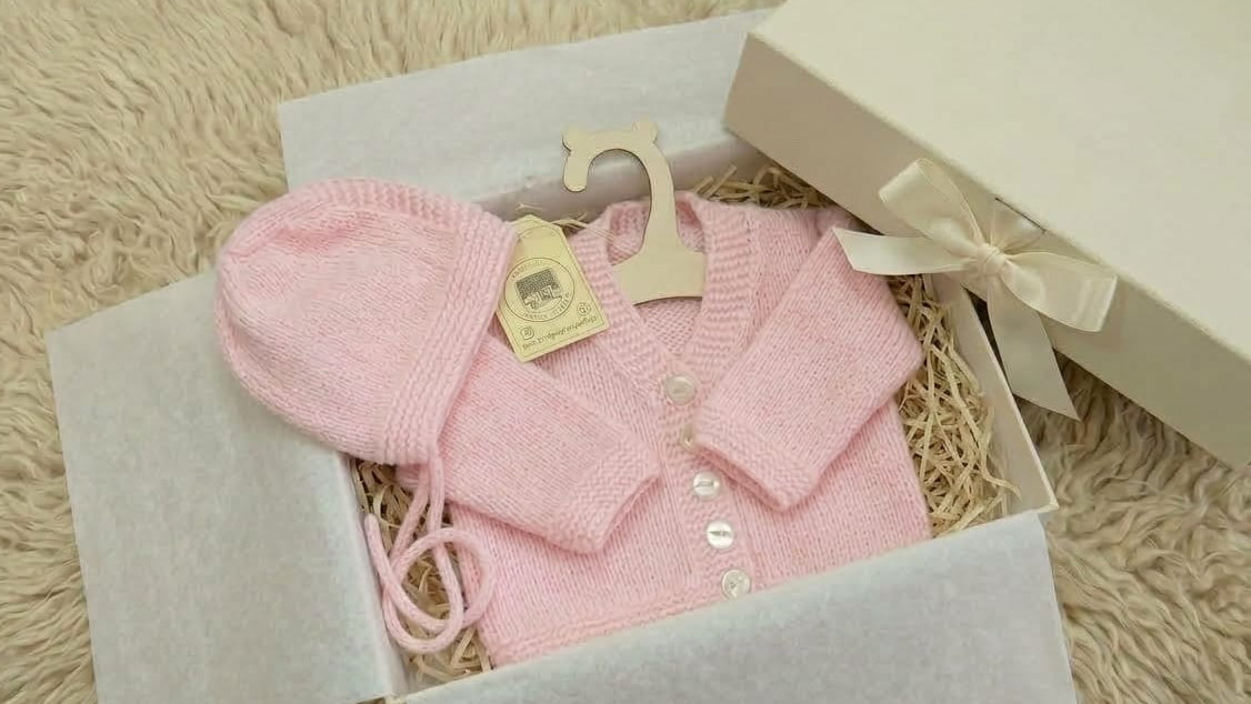 A light pink knitted baby cardigan and matching bonnet set displayed in an open gift box with tissue paper and shredded filler, alongside a cream-colored presentation box with a ribbon.