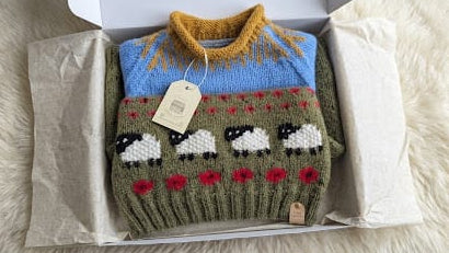 A folded hand-knit sweater displayed inside an open white gift box lined with tissue paper, set on a white faux fur surface. The sweater features a yellow and blue knit collar, a green body with a pattern of white sheep and red flowers, and a small wooden brand tag.