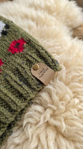 Close-up of a green knit fabric accessory with red and white floral embroidery, a tan leather tag reading 'SODA PEAK VALLEY', and a gold-tone snap, resting on a cream-colored shaggy rug.
