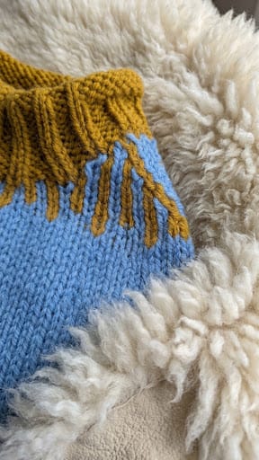 Close-up of a hand-knit sweater featuring a light blue body, a mustard yellow ribbed collar with matching fringe, and a thick white sherpa lining.