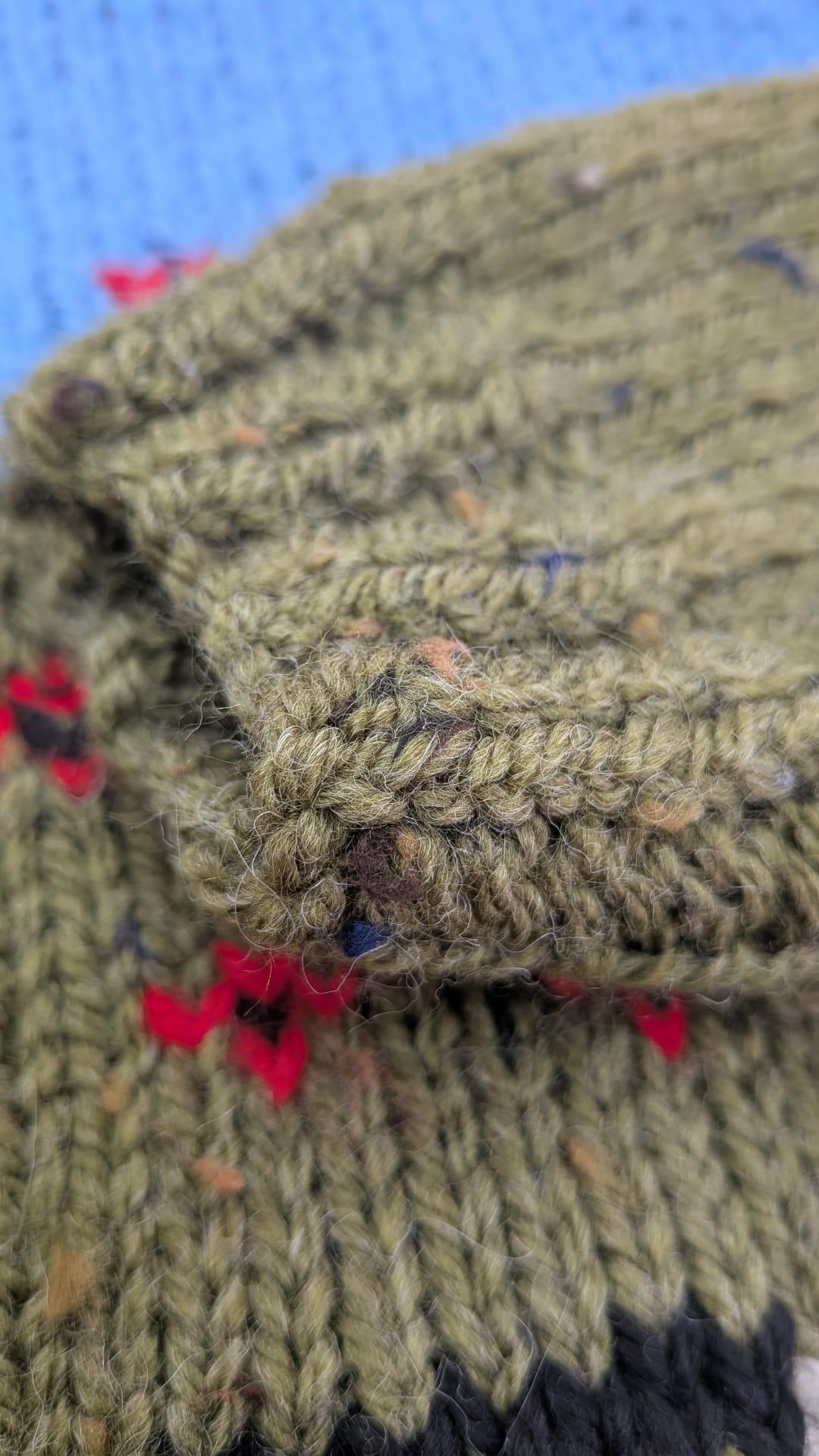 Close-up of a chunky knit beanie in olive green yarn with flecks of red, blue, and brown, featuring a folded cuff and a blue knitted background.