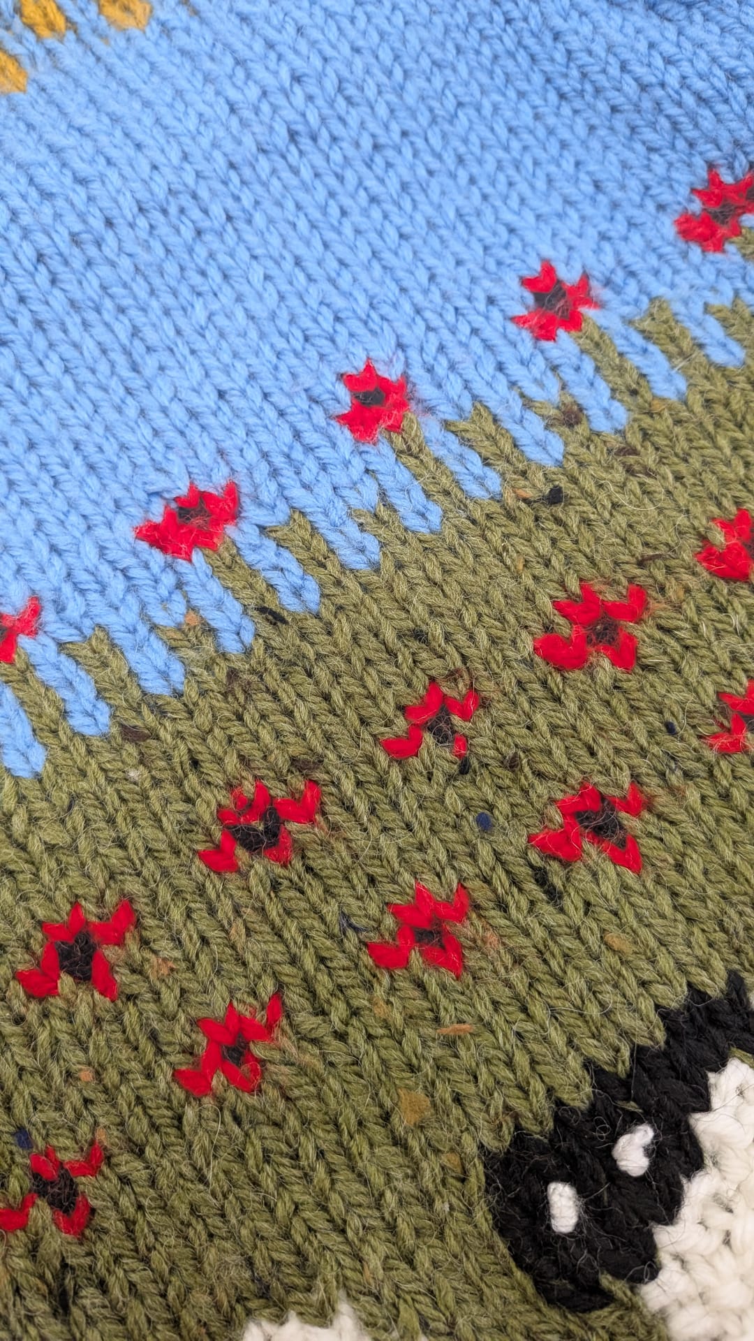 Close-up of a knitted textile with a blue upper section, an olive green lower section, and small red flowers with dark centers woven throughout.