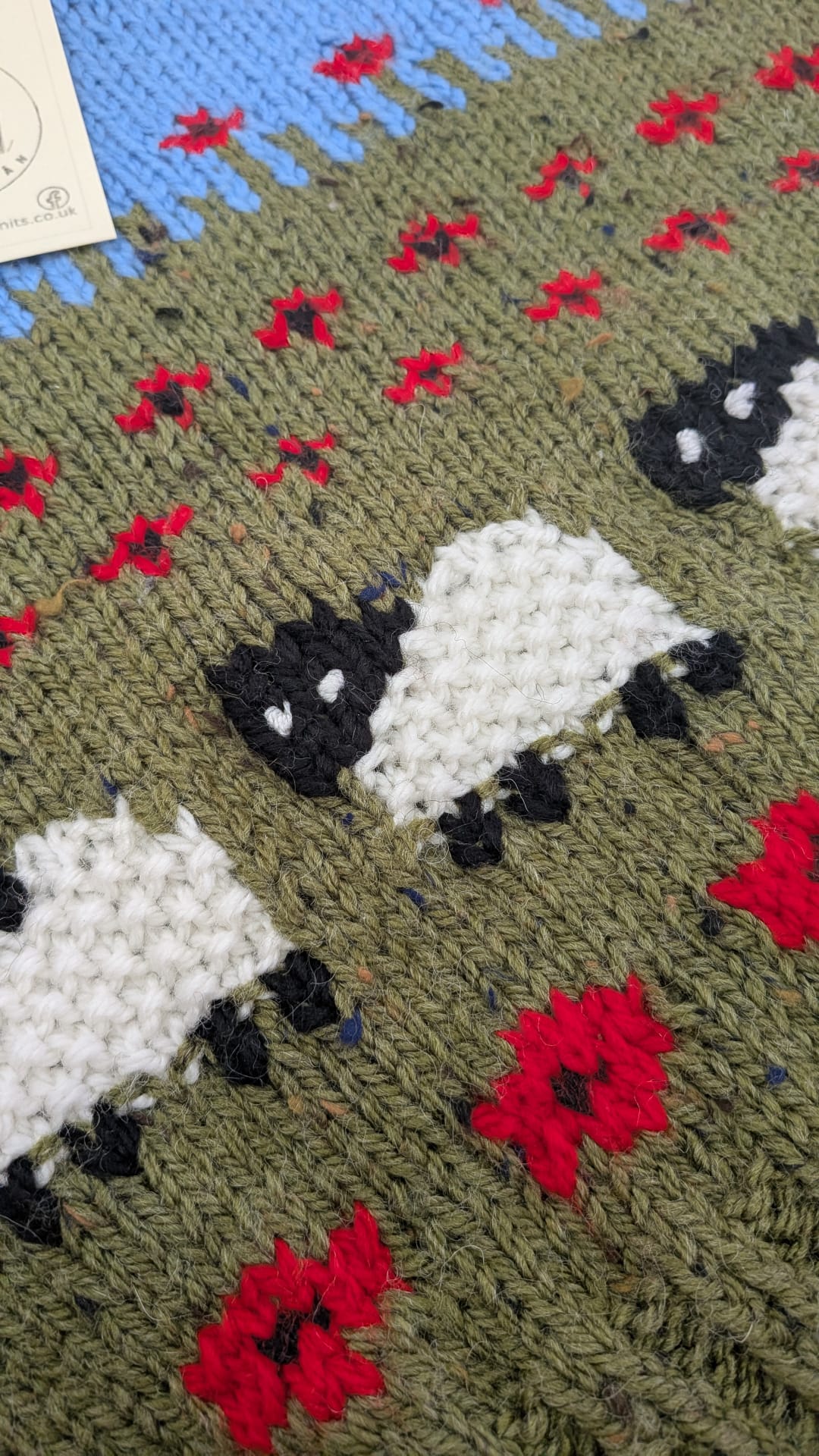 Close-up of a knitted blanket featuring sheep and red flowers on an olive green background, with a blue section and a small white tag visible in the top left.