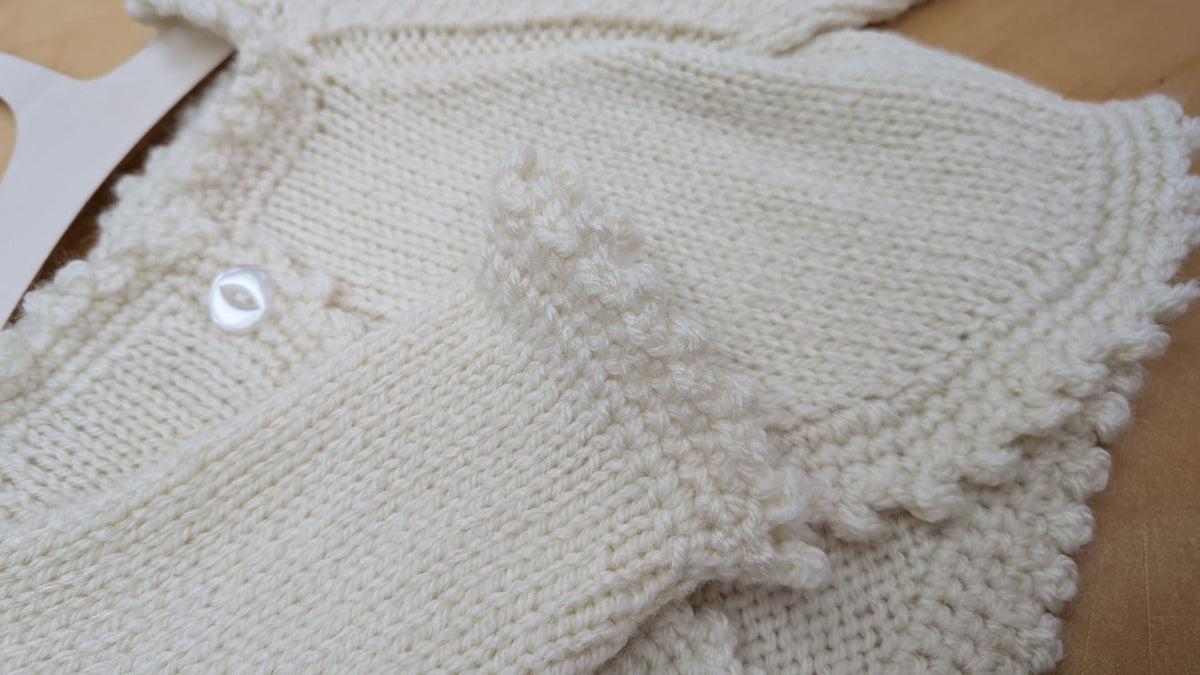Close-up of a cream-colored hand-knit baby cardigan with textured bobble stitches and a white button, laid flat on a light wood surface.
