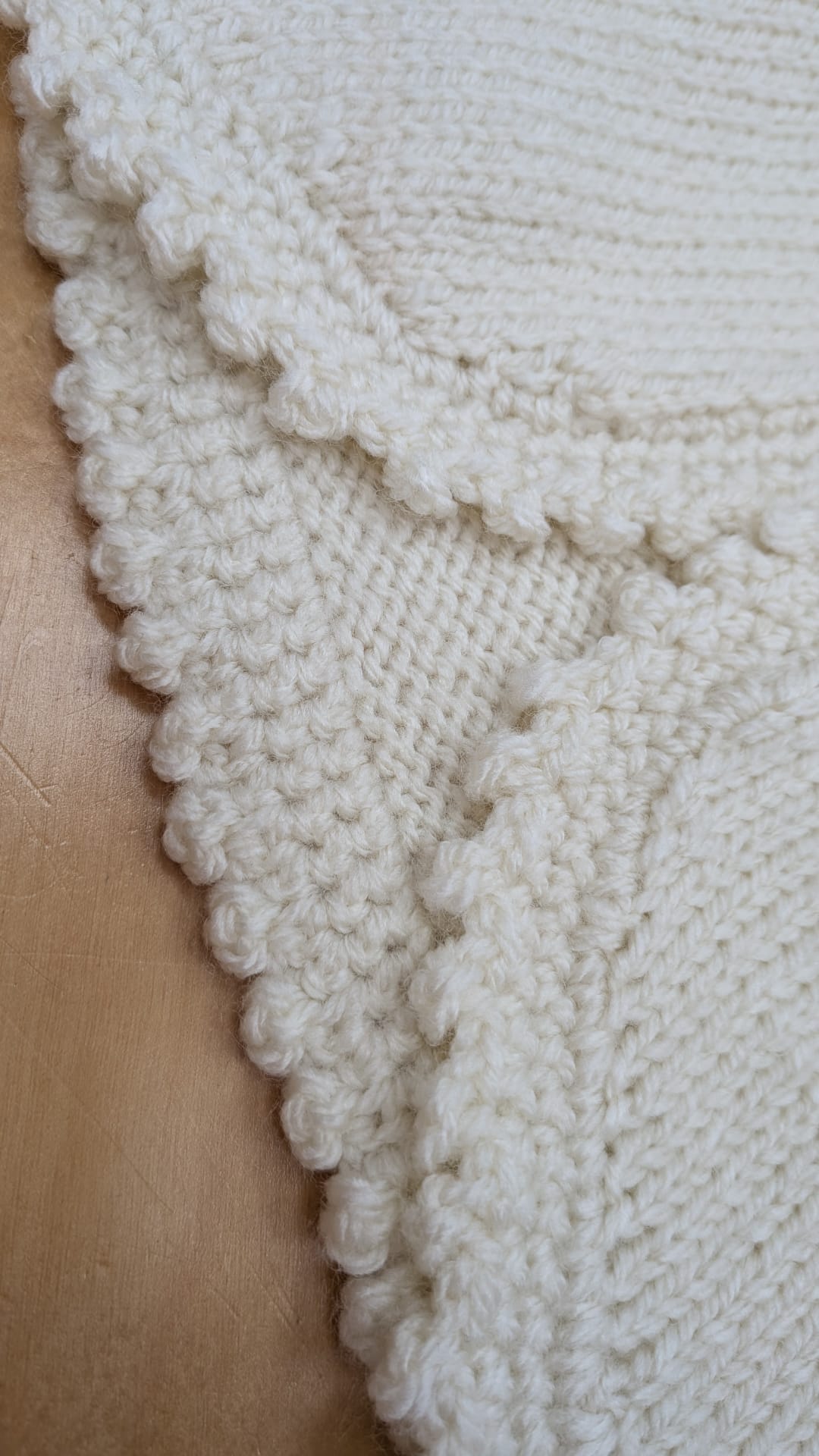 Close-up of a cream-colored knitted textile, likely a blanket or throw, showing textured knit patterns and a scalloped edge, laid over a light brown surface.
