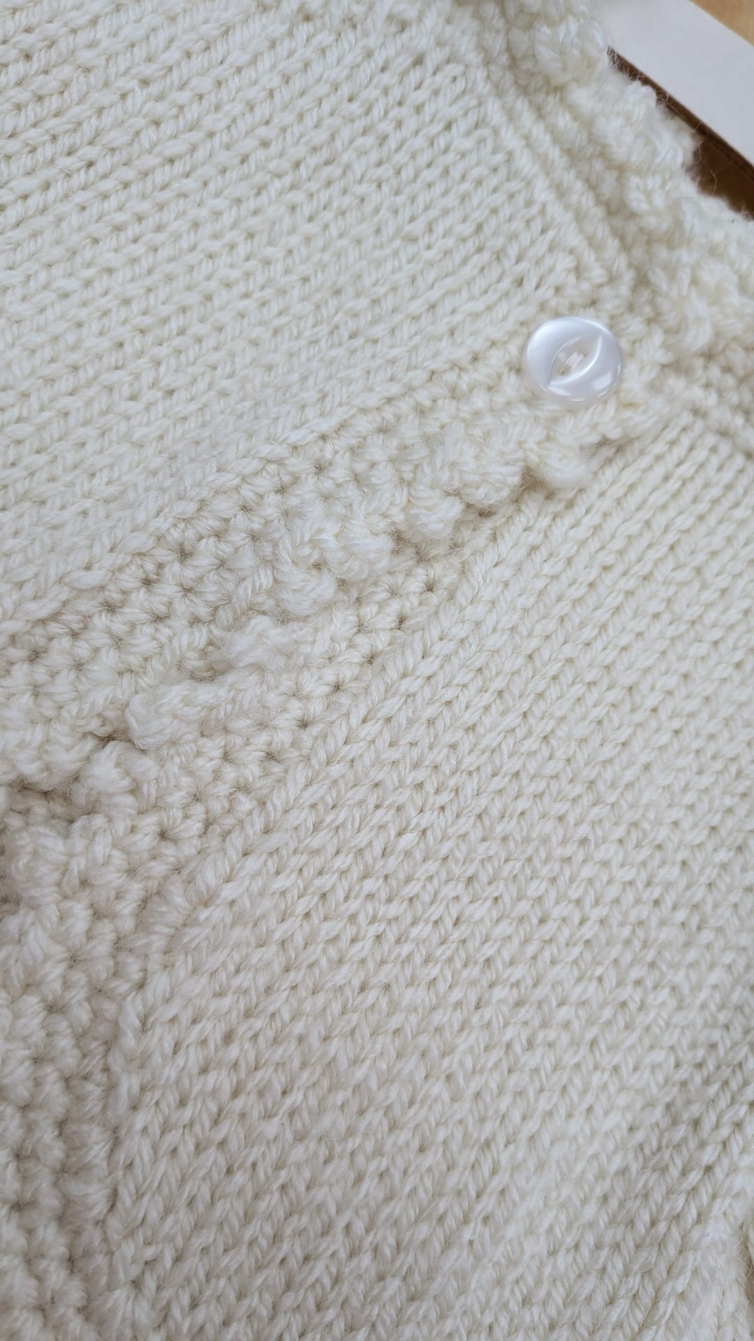 Close-up of a cream-colored knitted garment, showing textured knit patterns and a small white button.