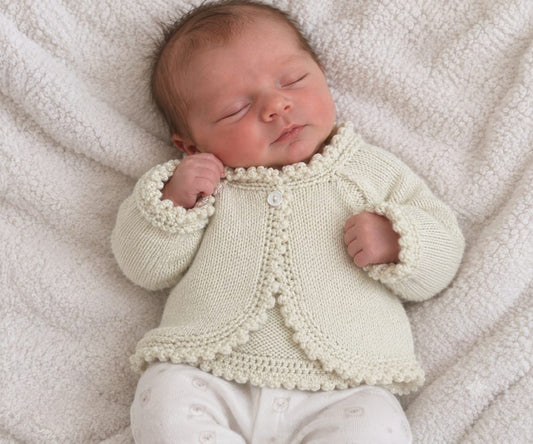 A sleeping newborn baby wearing a cream-colored hand-knit cardigan with pom-pom trim and white pants, lying on a soft textured cream blanket.
