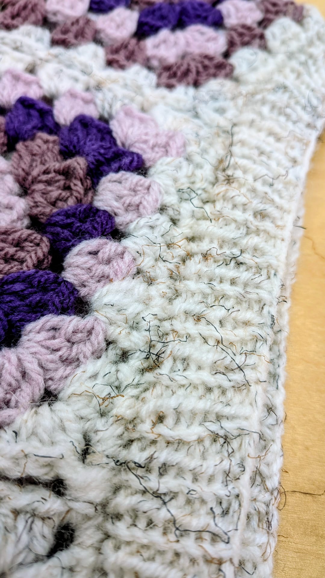 Close-up of a crocheted textile corner showing cream/off-white stitches, lavender and mauve floral sections, and dark purple accents, with loose fibers visible on a light wood surface.