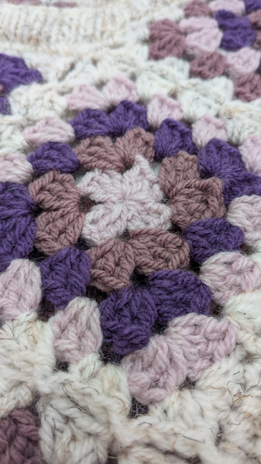 Close-up of a crocheted or knitted textile with a hexagonal granny-square pattern in cream, light pink, mauve, and purple yarn.