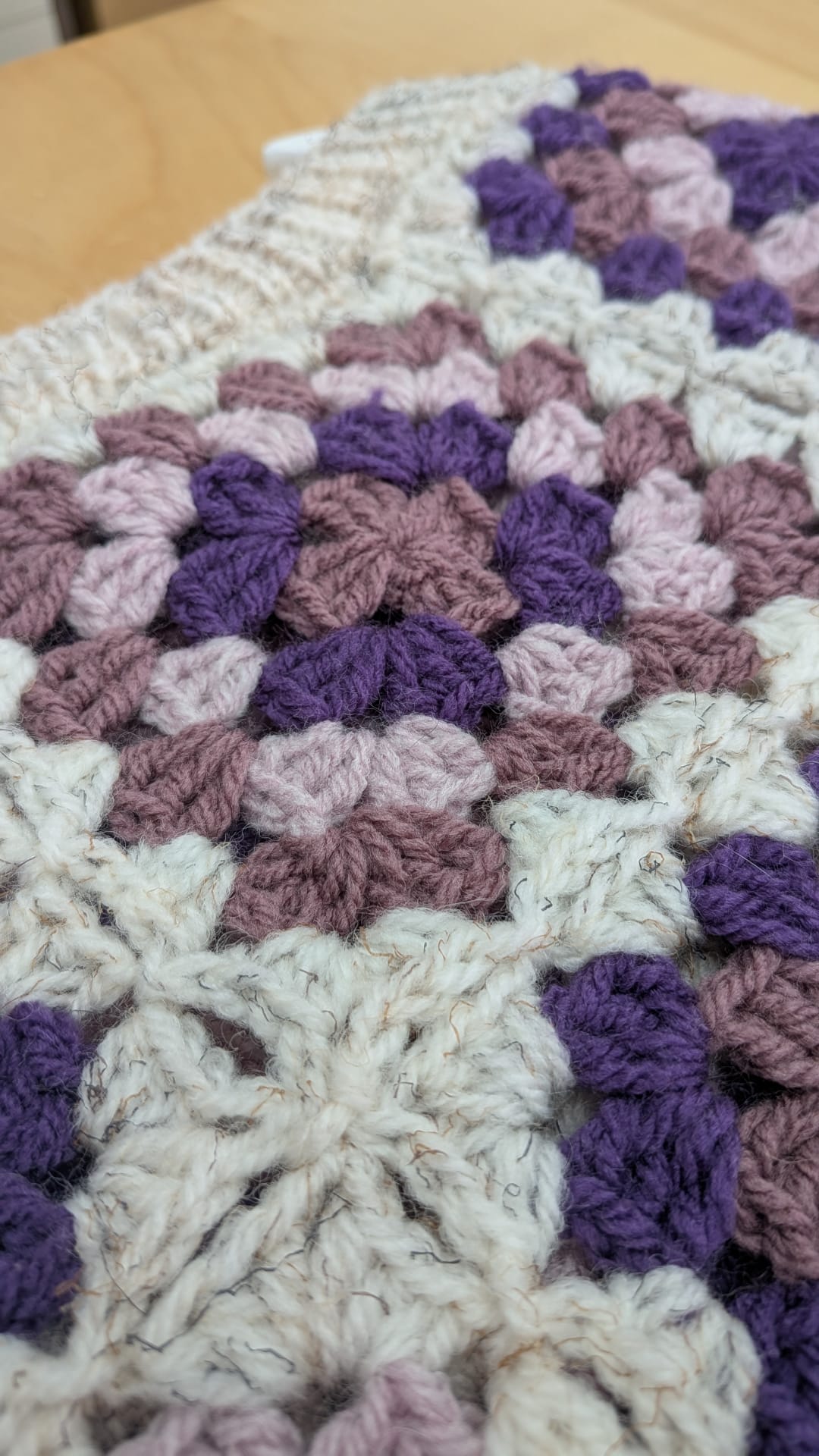A close-up of a handmade crochet granny square blanket featuring textured purple, dusty mauve, and cream yarn blocks on a light wood background.