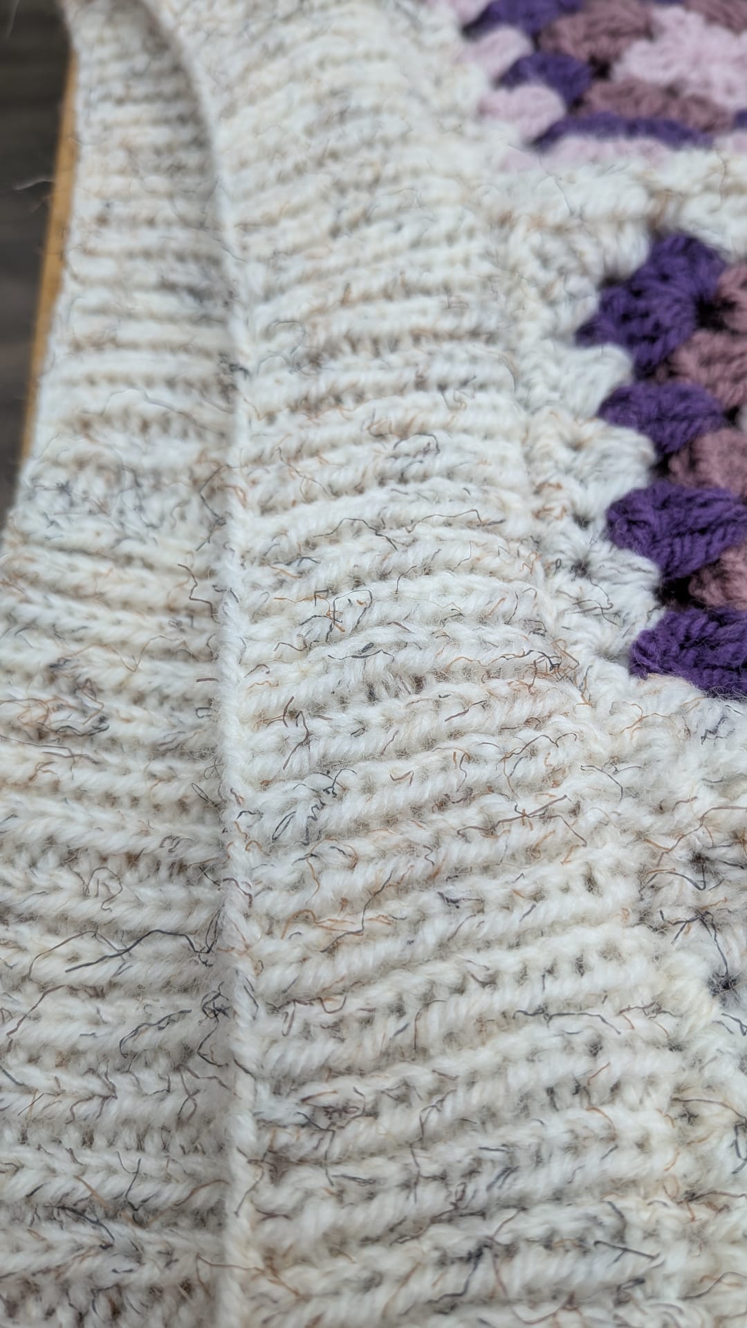 Close-up of a chunky knit or crocheted textile in a heathered off-white/cream color, with a row of purple and mauve floral or scalloped accents along one side.