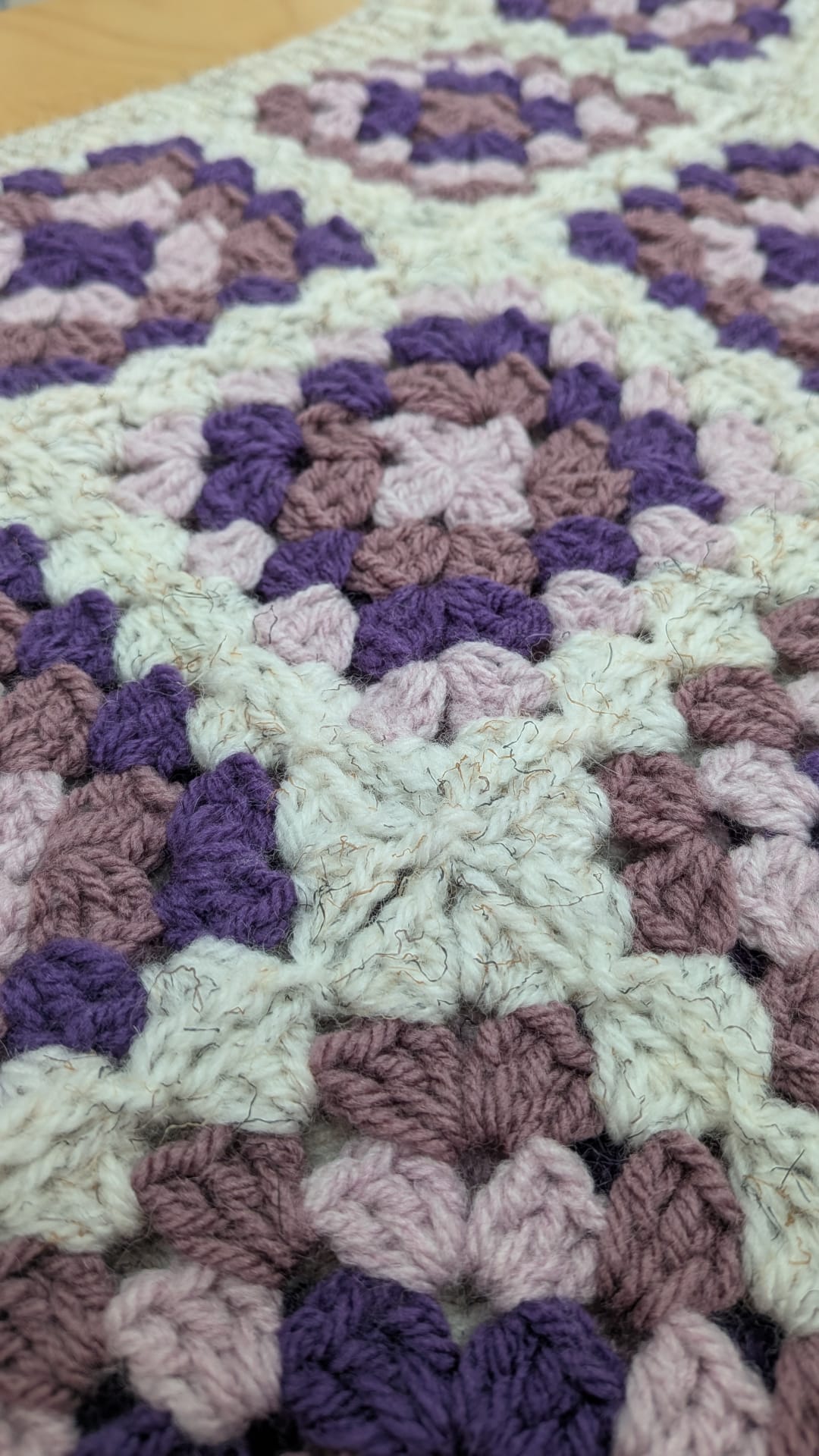 A close-up of a crocheted or knitted textile with repeating granny-square style motifs in cream, lavender, mauve, and dark purple.