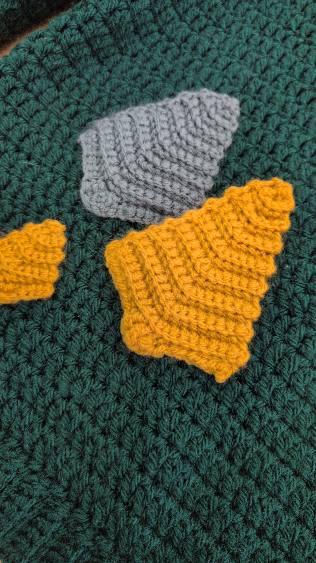 Close-up of a crocheted textile with a dark green textured base, a gray textured patch, and mustard yellow textured patches.
