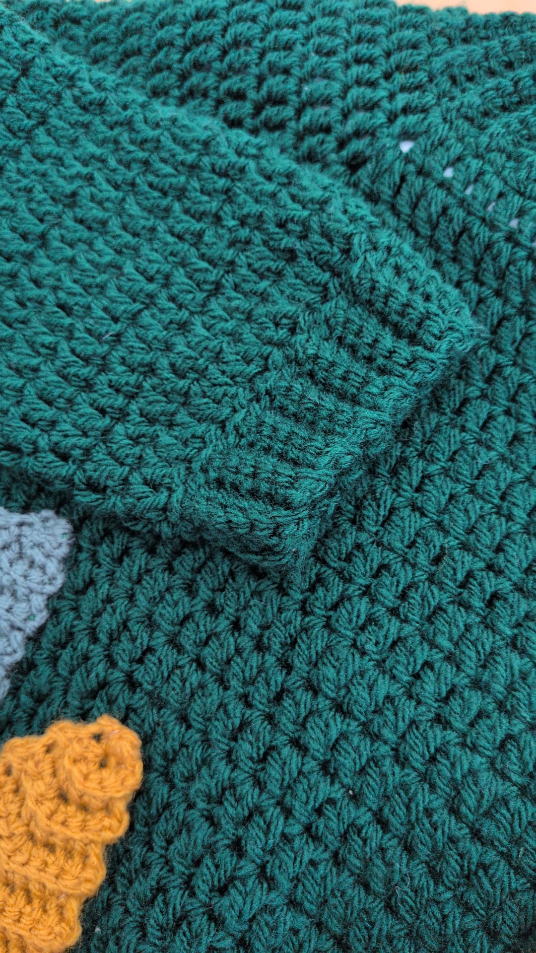 Close-up of a dark teal green crocheted textile, showing the detailed knit texture and a folded corner. A small portion of a mustard yellow crochet item is visible in the bottom-left corner.