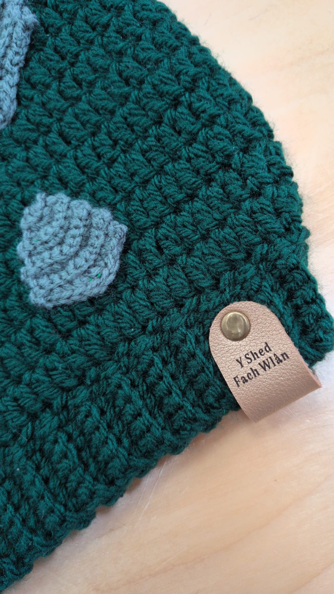 Close-up of a dark teal green crocheted beanie with a light blue-gray leaf applique and a tan leather tag reading 'Y Shed Fashions' on a light wood background.