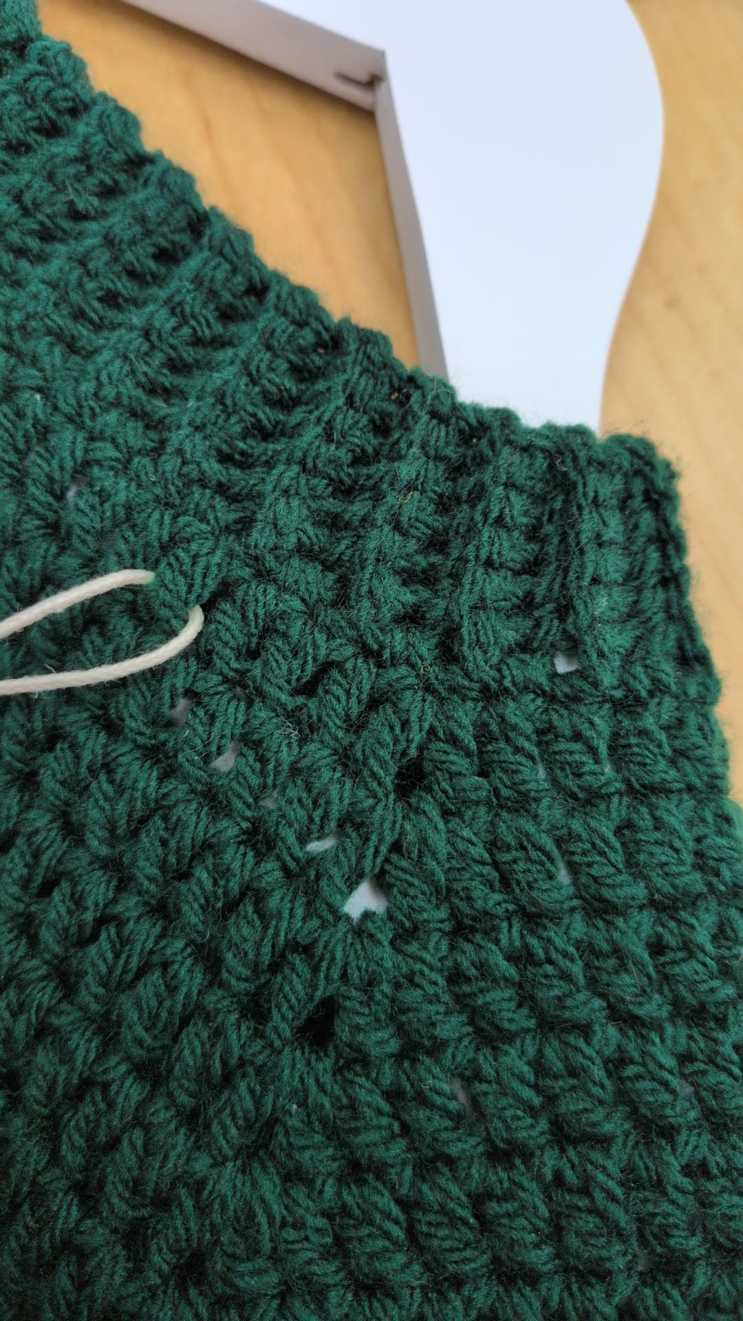 A close-up of a dark green crocheted fabric, showing the textured knit pattern and a white thread loop, resting on a light wood surface next to a white hanger.
