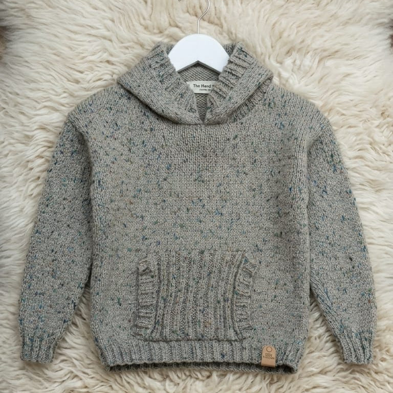 A gray knit hooded sweater with a speckled texture and ribbed front pocket, displayed on a white hanger over a fluffy cream faux-fur surface.