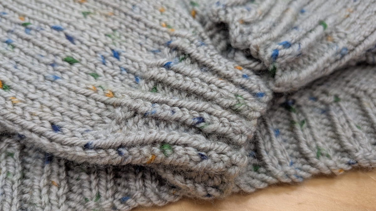 Close-up of a gray knitted garment, likely a sweater or scarf, featuring a variegated texture with small blue, green, and orange flecks, laid on a light wood surface.