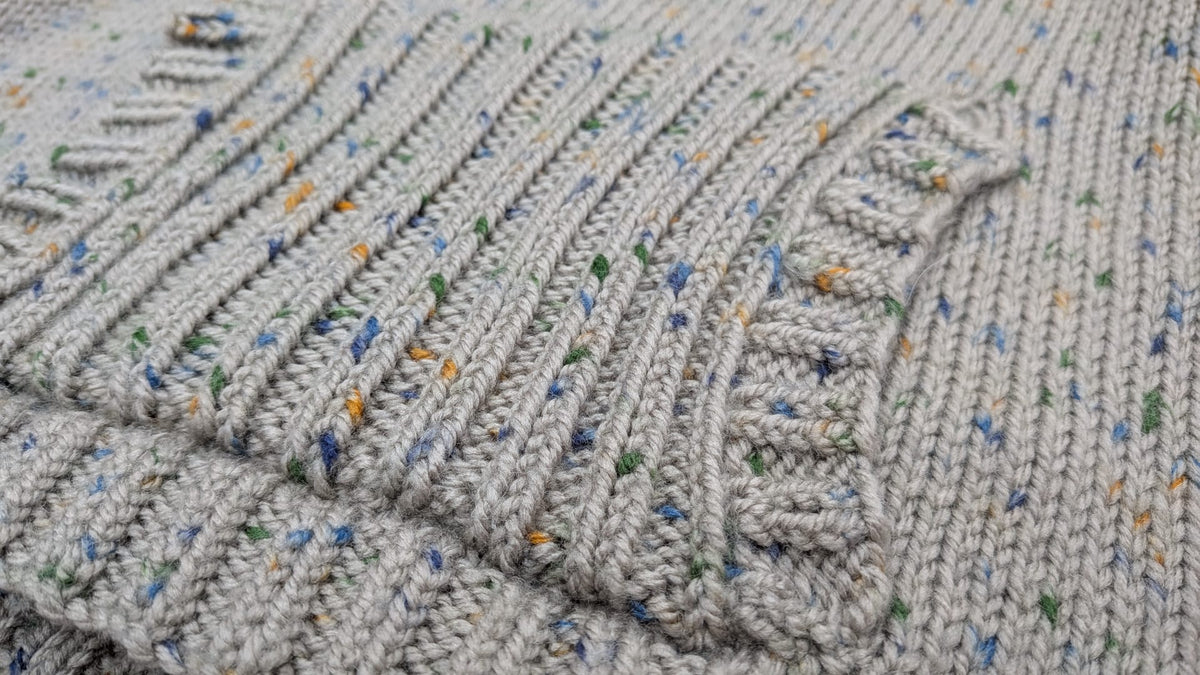 Close-up of a chunky knit blanket or throw in a light oatmeal gray color with small multicolored flecks of blue, green, and orange yarn.