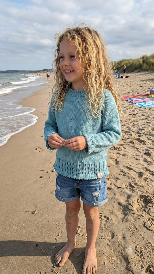 A young girl with long curly blonde hair stands on a sandy beach wearing a light blue knit sweater and denim shorts, looking to the side with a smile. The ocean waves and cloudy sky are in the background.