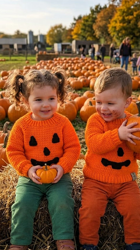 Two young children sitting on hay bales in a pumpkin patch, wearing matching bright orange knit sweaters with jack-o'-lantern faces. The girl holds a small pumpkin, and the boy holds a larger pumpkin.