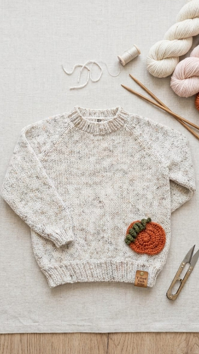 A cream-colored knit sweater with a small orange and green crocheted pumpkin patch on the lower right front, styled in a flat lay on a linen fabric background with yarn, knitting needles, scissors, and thread.