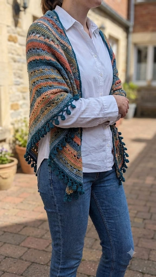 A person outdoors wearing a multicolored striped knit shawl with dark teal tassels over a white button-up shirt and blue jeans.