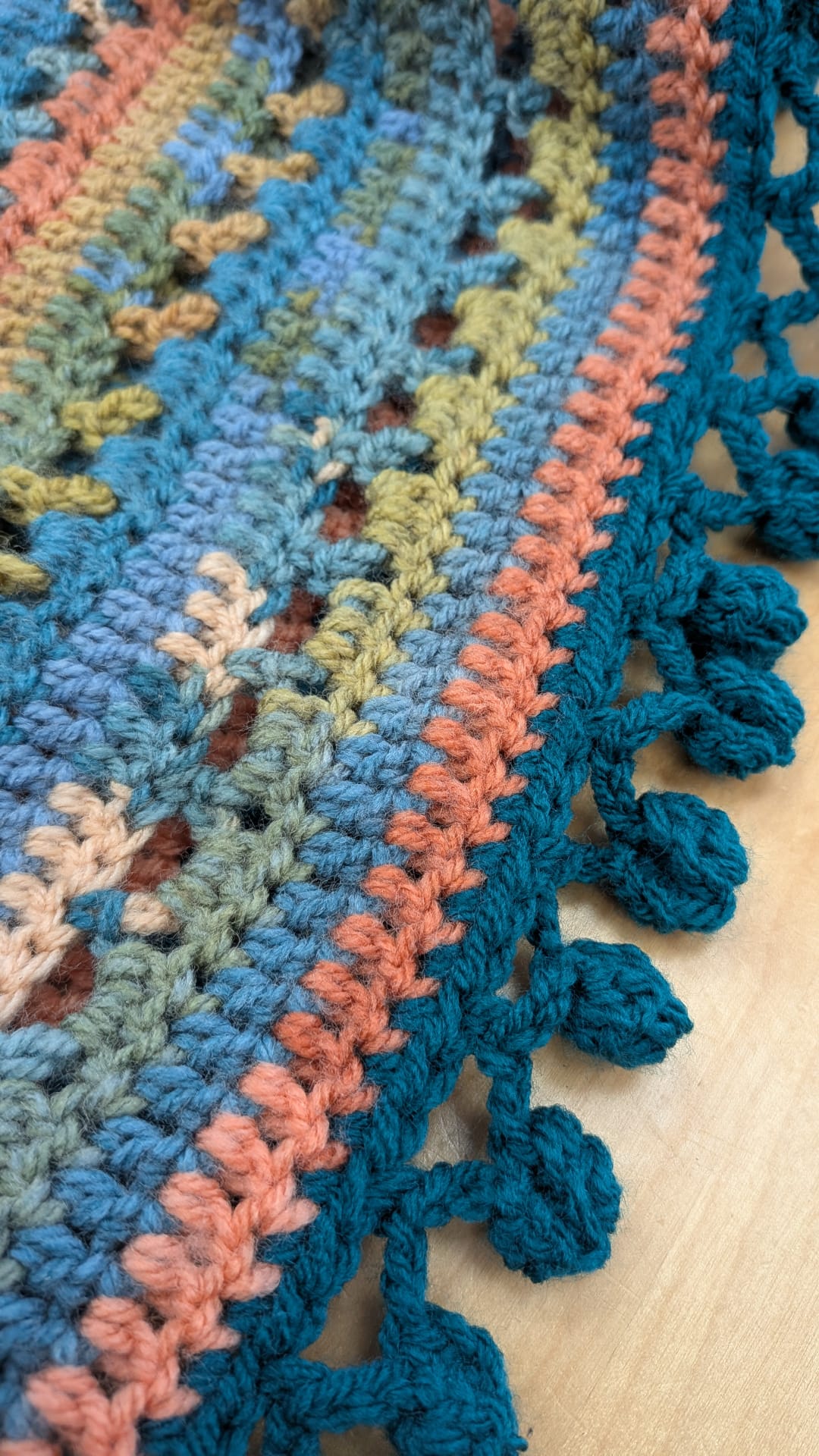 A close-up of a handmade crocheted textile, likely a blanket or scarf, featuring a multicolor pattern of blue, teal, peach, olive green, and brown yarn with a decorative pom-pom border.