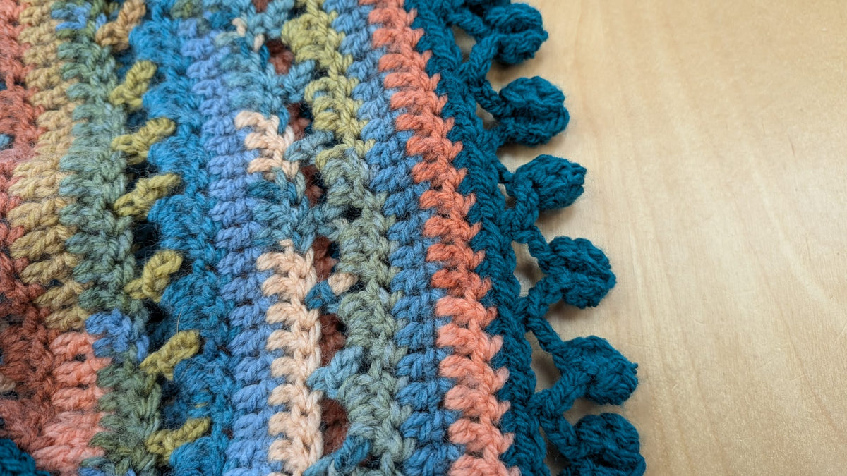 Close-up of a crocheted textile with multicolored stripes in blue, teal, peach, beige, and olive, finished with a tassel trim, laid on a light wood surface.
