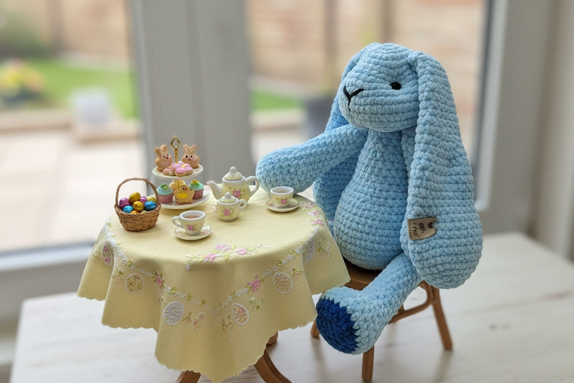 Blue crochet bunny sitting at a small table with tea set in a home setting
