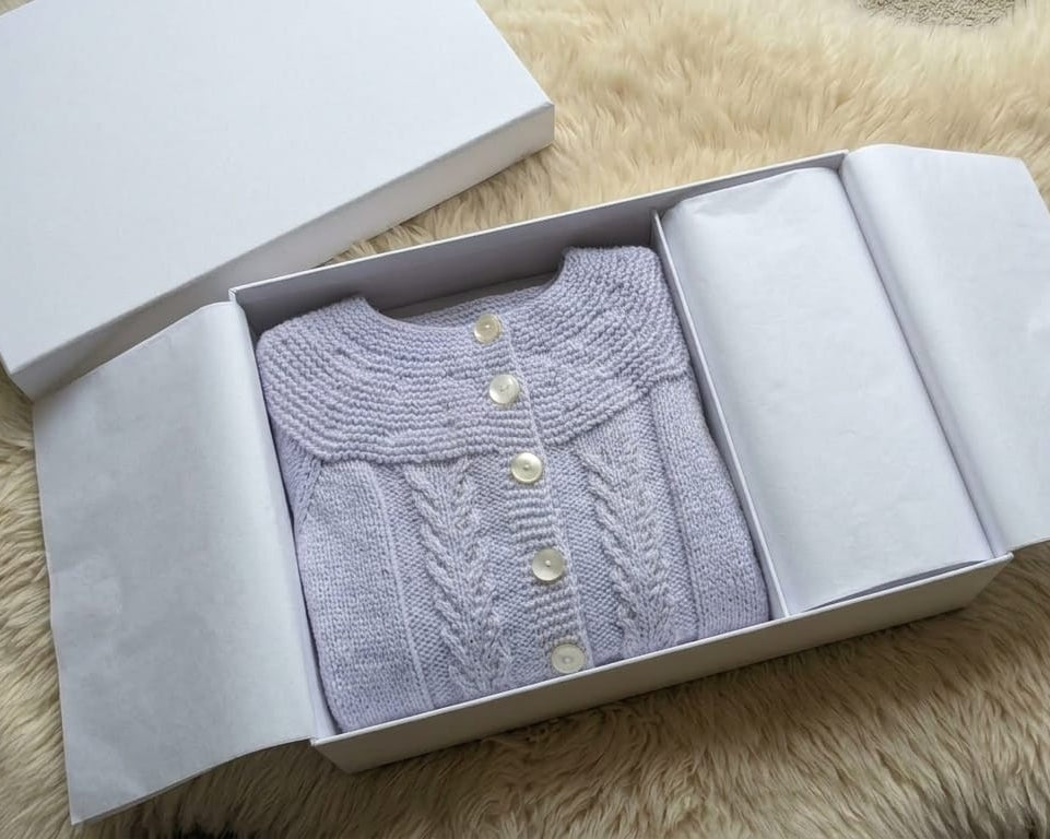 A light lavender knit cardigan with cream buttons, neatly folded inside an open white gift box lined with white tissue paper, set on a beige faux fur surface.