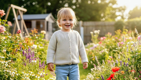 A young child stands smiling in a sunlit garden, wearing a cream knit sweater, light blue shorts, and white sneakers, surrounded by colorful flowers and greenery.