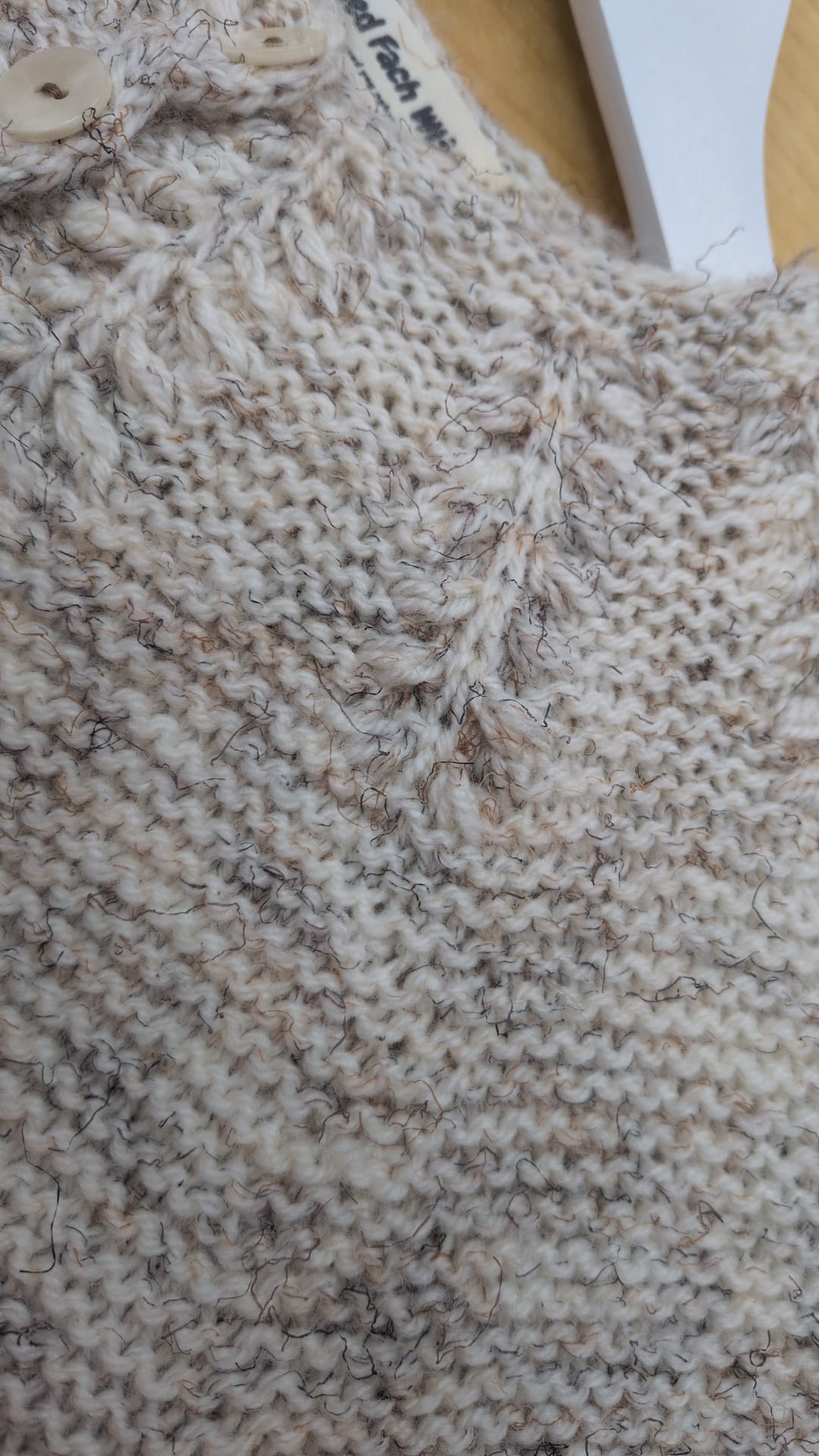 Close-up of a chunky knit sweater in a heathered cream and beige yarn, showing the detailed knit texture and a small portion of the wooden buttons at the collar.
