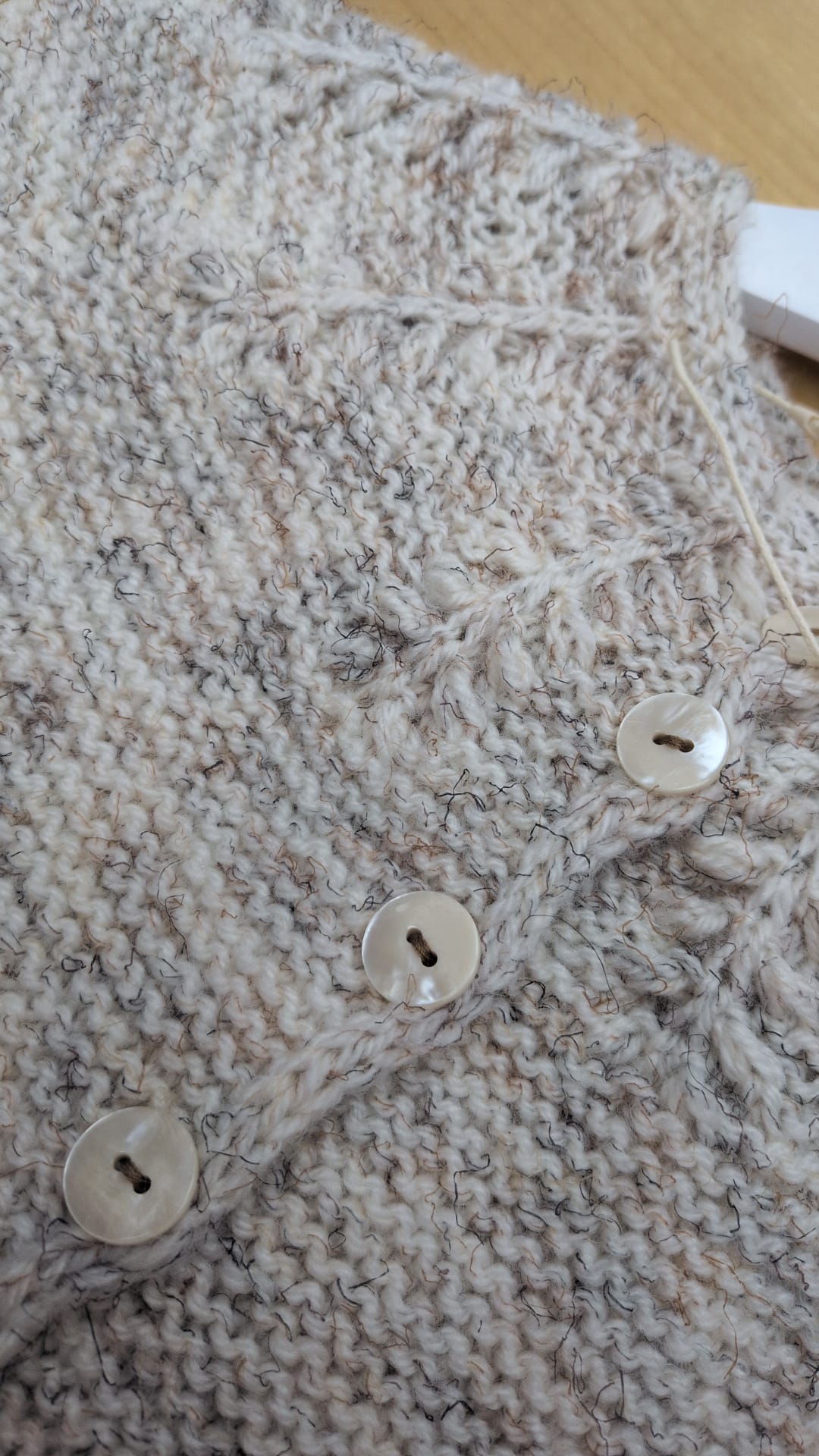 Close-up of a chunky knit cardigan in a heathered oatmeal beige and gray yarn, featuring three large white buttons down the front.