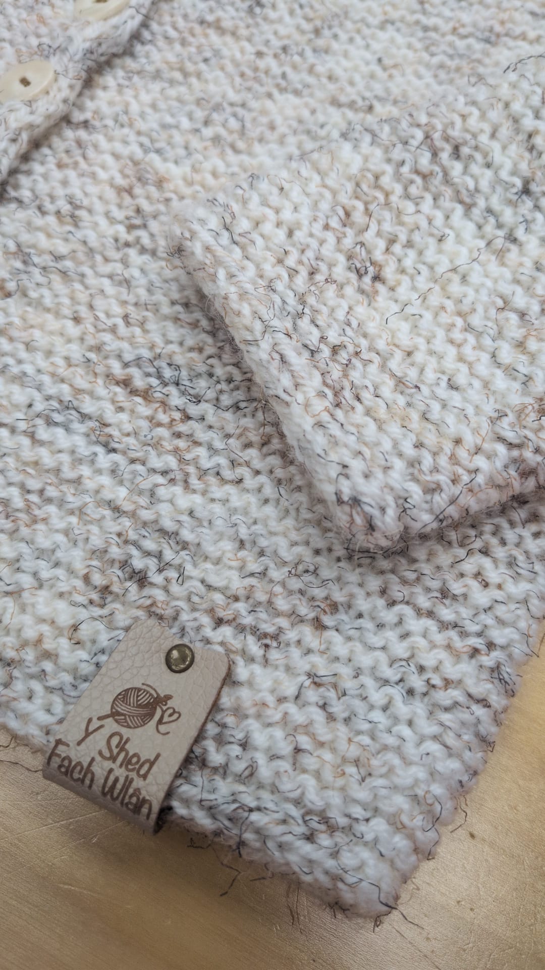 Close-up of a chunky knit cardigan in a cream and beige variegated yarn, featuring a folded cuff and a leather brand tag that reads 'Y Shed Faux Wool'.
