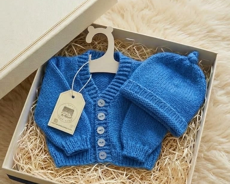 A blue knitted baby cardigan and matching hat presented in a gift box with shredded paper, a cream-colored box, a navy ribbon, and a small card on a cream faux-fur surface.