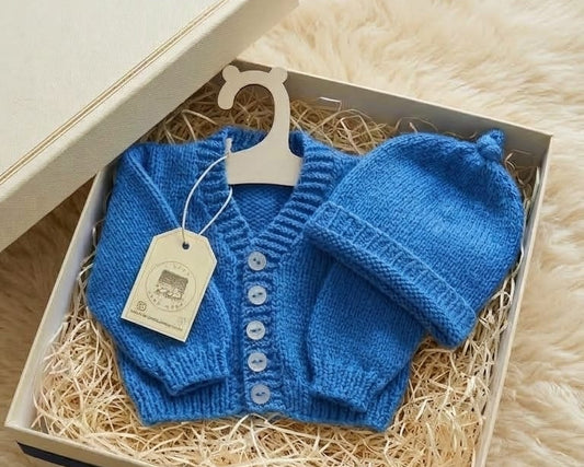 A blue knitted baby cardigan and matching hat presented in a gift box with shredded paper, a cream-colored box, a navy ribbon, and a small card on a cream faux-fur surface.