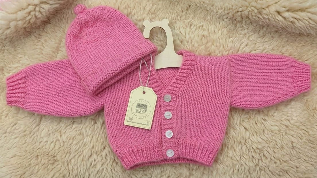 A pink knit baby clothing set including a hat, booties, and a small sweater, laid flat on a beige faux fur surface. The set has a cream paper hangtag attached.